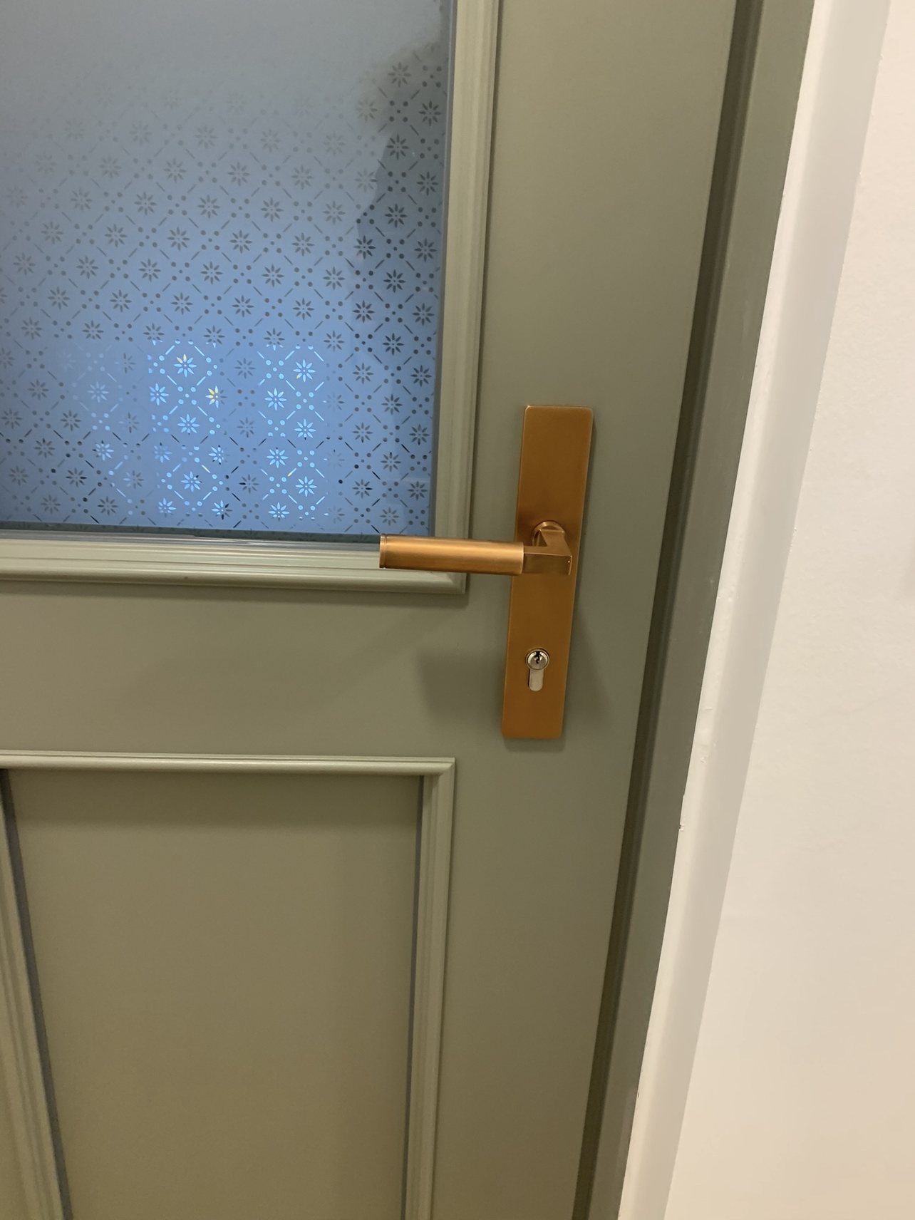 Nahaufnahme der Wohnungseingangstür: Ornamentglas mit Rautenmuster und Sternen, dazu bronzefarbener Langschildbeschlag mit Drücker und Profilzylinder