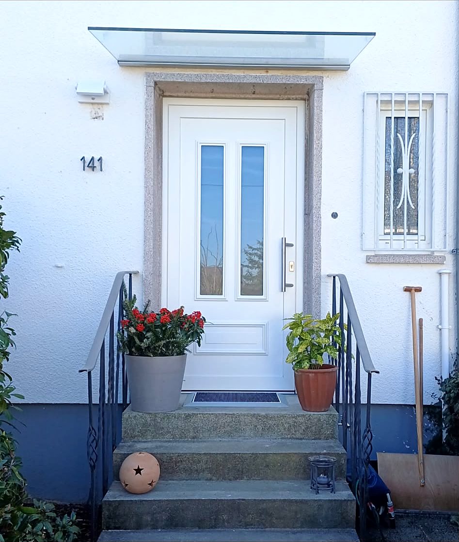 Weiße Haustür mit hohem Glasausschnitt und Vordach, davor eine Treppe mit Blumenkübeln