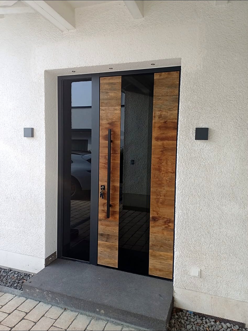 Graue Haustür mit senkrechtem Holzdekor-Feld, langer Griffstange und Glasseitenteil